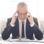 Handsome tired mature businessman in classic suit is massaging his temples while sitting in his office